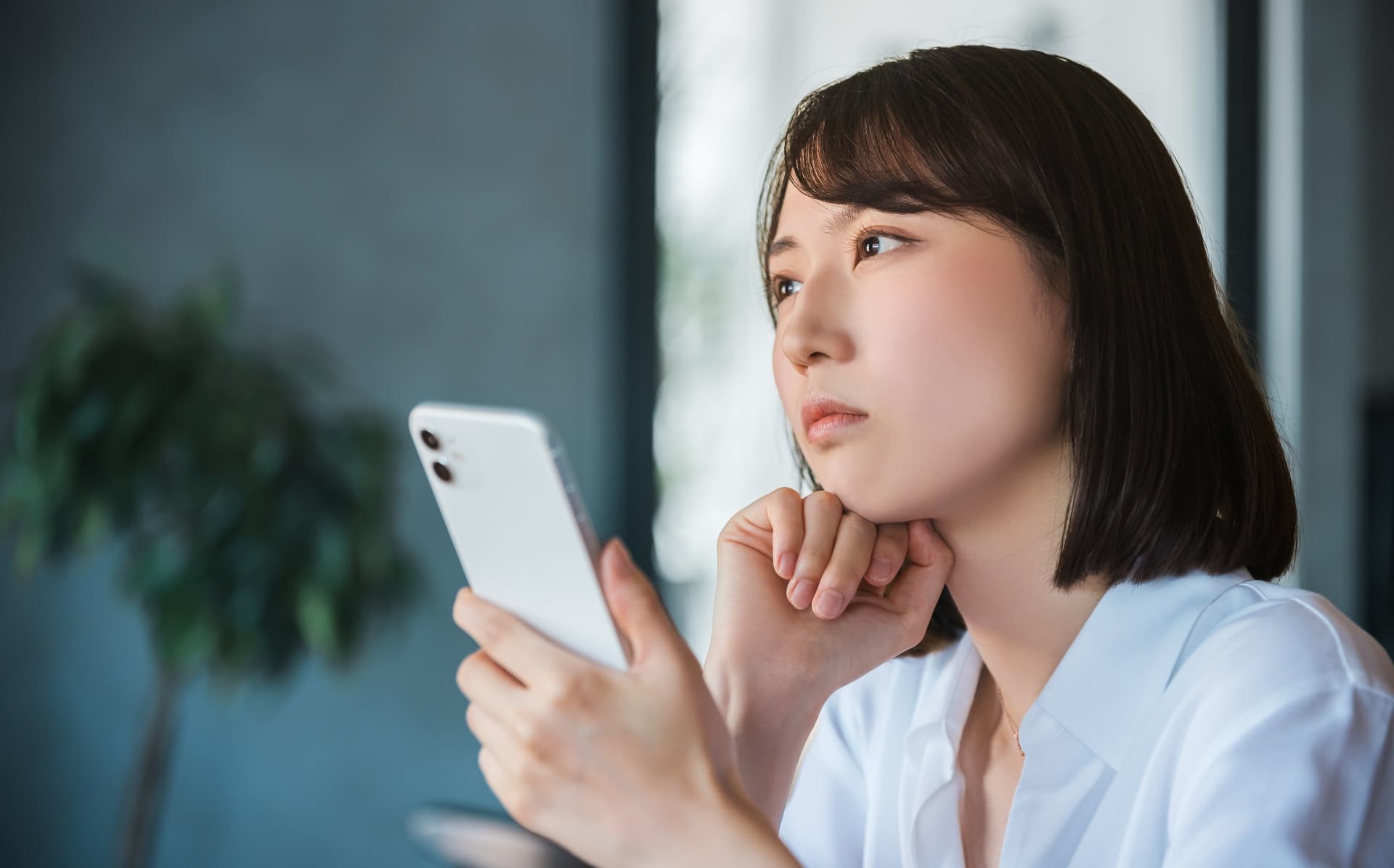 スマートフォンを片手に、顎に手を当てて何かを深く考え込んでいる様子の女性の写真