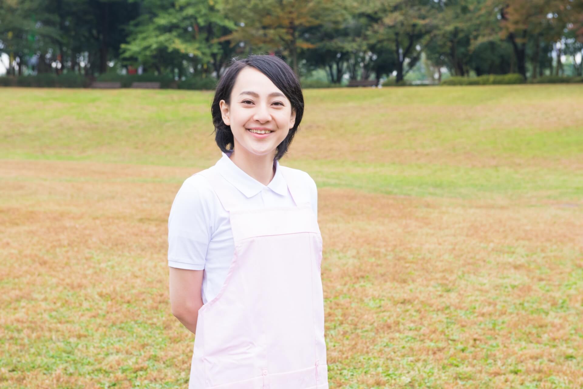 看護服を着用した女性スタッフが、外の芝生の上で笑顔でこちらを見つめて立っている写真