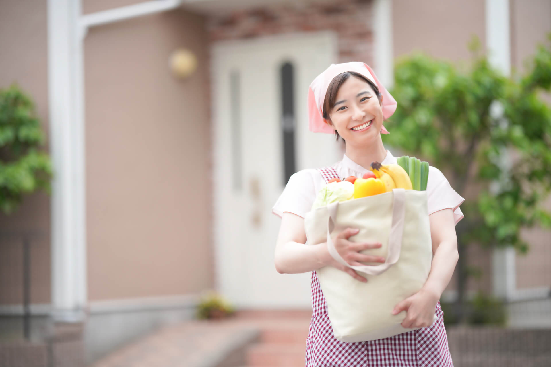エプロンと帽子を身につけた女性家政婦スタッフが、家の前でたくさんの食材が入った買い物袋を抱えて、笑顔でこちらを見つめている写真