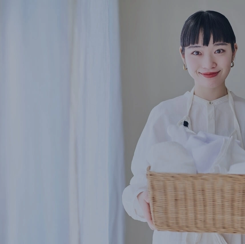 洗濯物の入ったカゴを持ちながら笑顔でこちらを見つめる女性家政婦の写真