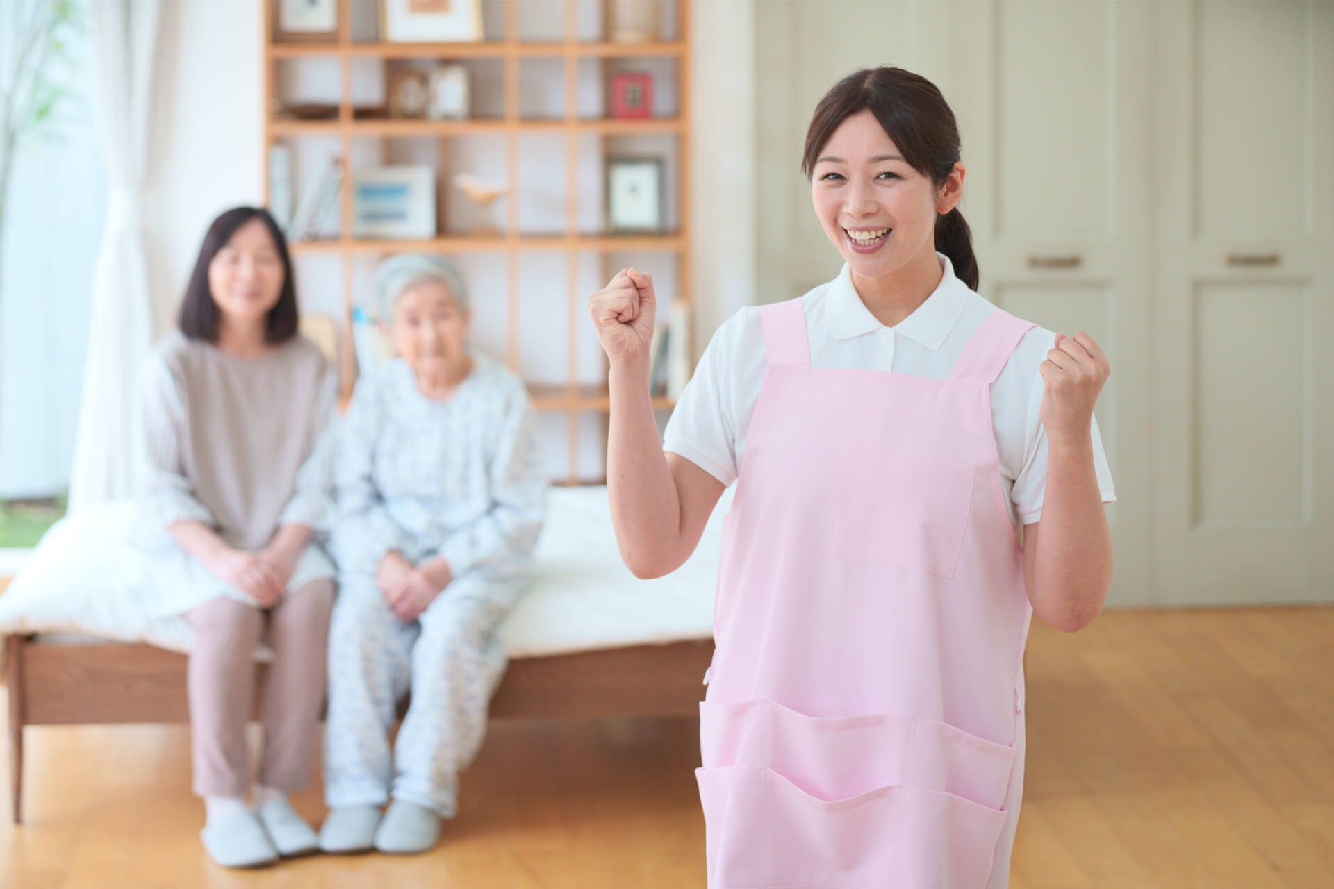 ガッツポーズをしながら笑顔でこちらを見つめる女性介護スタッフと、その背後でご高齢女性とご家族がベッドに腰掛けている写真