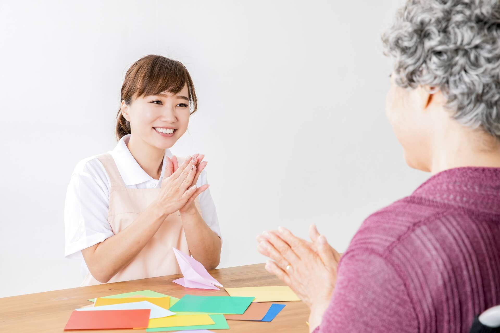 エプロンを身につけた女性介護スタッフが、車椅子に座った白髪のご高齢女性と笑顔で折り紙を楽しみ、指先の運動をしている写真